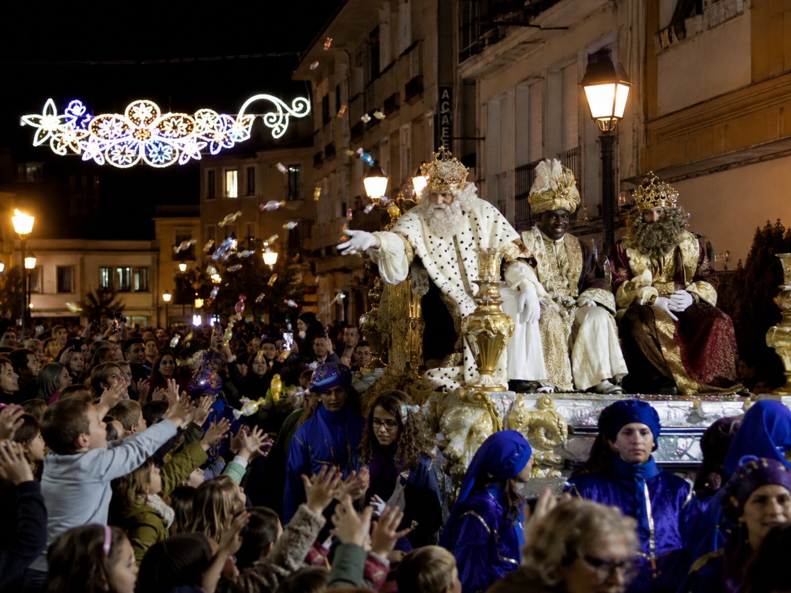 La cabalgata de los Reyes Magos — the Three Kings parade in Spain at night, with crowds of children reaching up to catch sweets